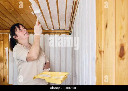Rolling paint, female decorator covering timber ceiling with white coating to refresh wooden finish and enhance room illumination. Stock Photo