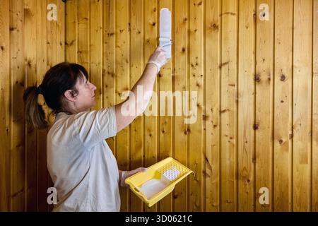 Woman softly applies fresh coat of paint to wooden wall panel using paint roller, standing with paint tray in left hand, showing precise paint Stock Photo