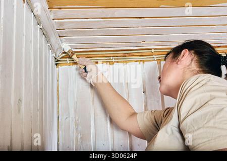 Coating edge, female painter applying white layer along meeting line of vertical and horizontal wood paneling to achieve consistent interior finish. Stock Photo
