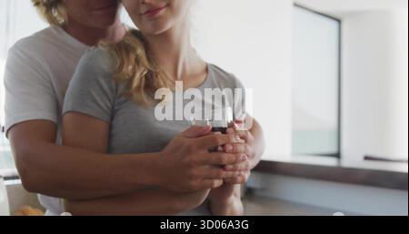 Hugging couple in casual T-shirts leaning in modern kitchen, with clear glass mug and countertop Stock Photo