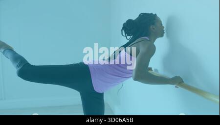 Practicing woman extending leg at barre in dance studio, wearing pink tank top and black leggings Stock Photo
