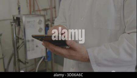 Lab technician in lab coat operating touchscreen tablet in control room, with gauges, pipes, wiring Stock Photo