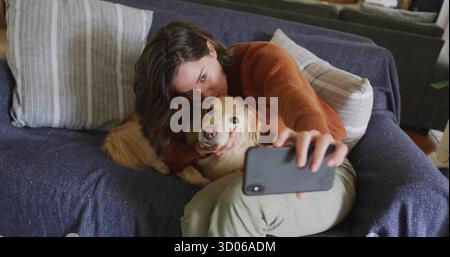 Reclining woman hugging retriever and taking selfie with phone on blue throw sofa, striped cushions Stock Photo