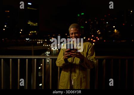 Woman smiling using phone on urban night balcony Stock Photo