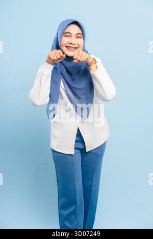 Portrait of a cheerful young Asian Muslim woman raising fists in a victorious or excited pose, isolated on a blue background. Stock Photo