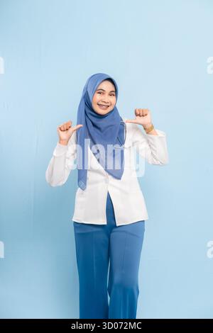 Portrait of a cheerful Asian Muslim doctor or nurse pointing thumbs to self, expressing confidence, isolated on a blue background. Stock Photo