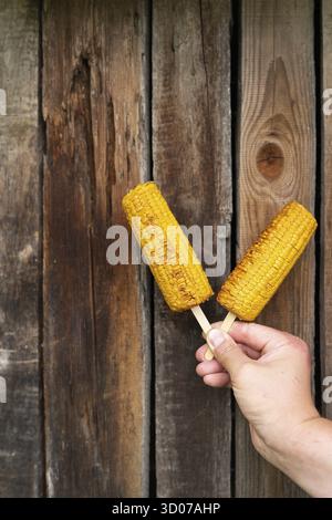In his hands, a man holds fried sweet corn with salt and butter on a stick on a grill against the background of a wooden old wall. Summer vegan dinner Stock Photo