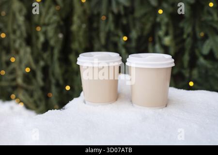 Two plastic cups stand on the snow against the backdrop of a green wall of a Christmas tree and bokeh from a garland. Coffee to go, a walk in the fres Stock Photo