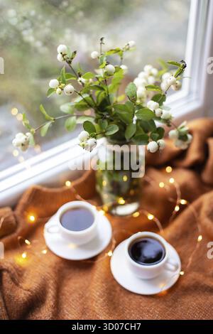 A white snowberry in a bouquet stands with two cups of coffee on the windowsill near the window, beautiful lights, bokeh in the window Stock Photo