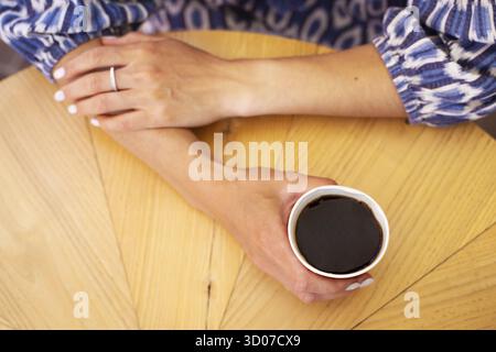 A wooden table on which there is a red plastic cup with coffee. The girl's hands are holding a glass of coffee Stock Photo