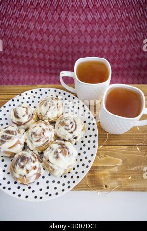 Two cups of black tea stand on a wooden tray. Fresh and fragrant cinnamon rolls close up lie on a plate with polka dots, beautiful morning. Close-up. Stock Photo