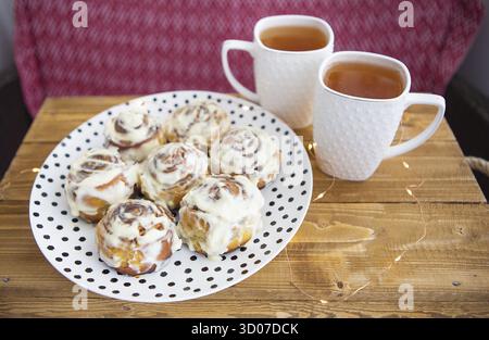 Two cups of black tea stand on a wooden tray. Fresh and fragrant cinnamon rolls close up lie on a plate with polka dots, beautiful morning. Close-up. Stock Photo