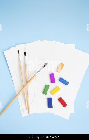 Multi-colored paints, watercolor brushes of different sizes lie together with watercolor paper on a blue background. Drawing lessons. View from above Stock Photo