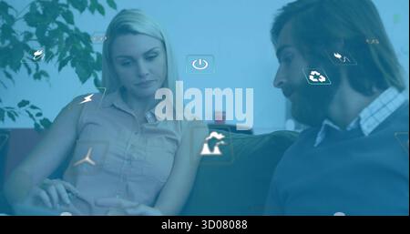 Coworkers discussing floating icons in office lounge on sofa near potted plant, with red coffee cup Stock Photo