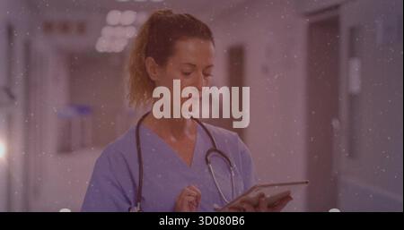 Nurse checking records on tablet in hospital corridor, with stethoscope, light-blue scrubs Stock Photo