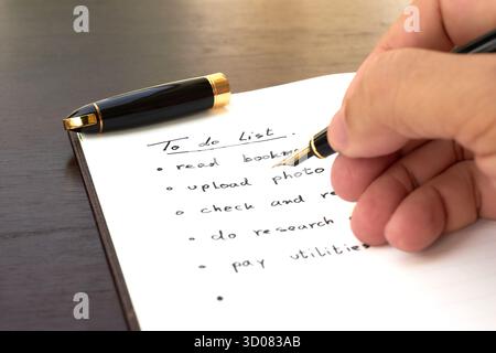 A hand holds a pen above a notebook filled with a to-do list. The list includes tasks like reading, uploading photos, and paying utilities in a simple Stock Photo