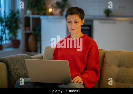 Tranquil mature woman work calmly on laptop on sofa in comfortable living room. Female productivity Stock Photo