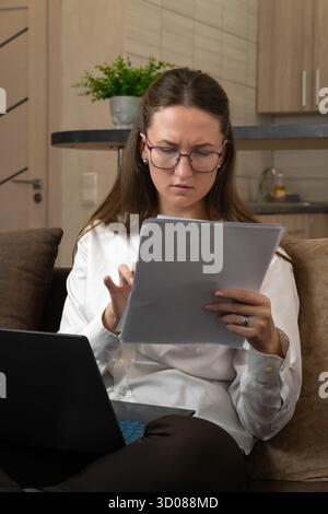 Focused businesswoman wearing glasses reviews important documents while working remotely on her laptop from the comfort of her home office, demonstrat Stock Photo