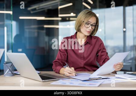 Businesswoman in a modern office desk, focused on analyzing financial reports and charts while writing notes, reviewing paperwork and planning strategy on a laptop Stock Photo
