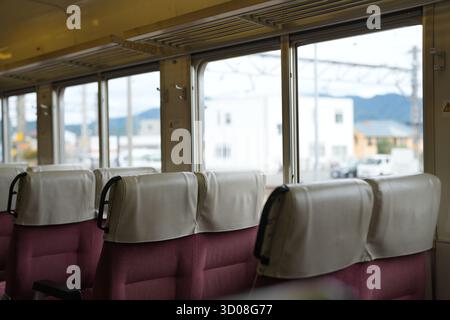 The train has a lot of empty seats. The seats are covered in leather and are facing the window. The train is on a track and is going through a city Stock Photo