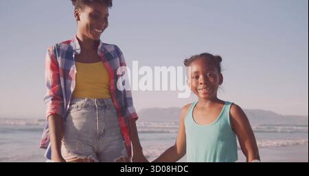 Walking mother and daughter holding hands wearing bright outfits on beach shoreline under clear sky Stock Photo