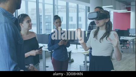 Gesturing woman using VR headset in windowed office, colleagues holding devices, copy space Stock Photo