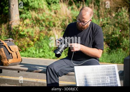 Bearded male photographer sits on bench, connecting camera to solar panel for charging. Integration of renewable energy into photography, using solar power to charge equipment during outdoor shoots. Stock Photo