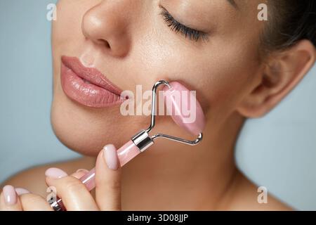 Close up photo of a young woman looking relaxed and smiling while using a natural rose quartz face roller. Face massage. Stock Photo