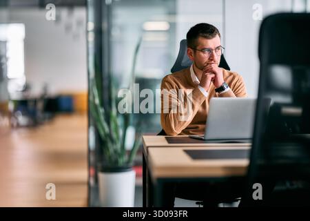 Focused businessman sitting at desk with laptop, thoughtfully analyzing data or planning strategy in a modern office environment, representing Stock Photo