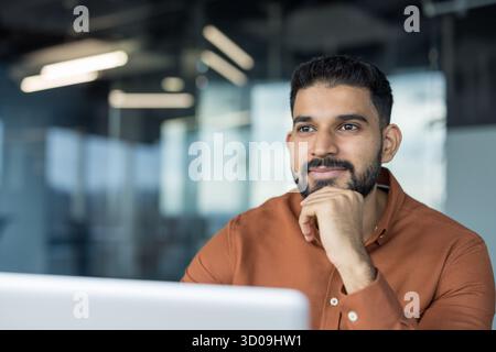 Young indian businessman contemplating future business ideas, sitting at his desk with a thoughtful expression and hand on chin in a modern office environment Stock Photo