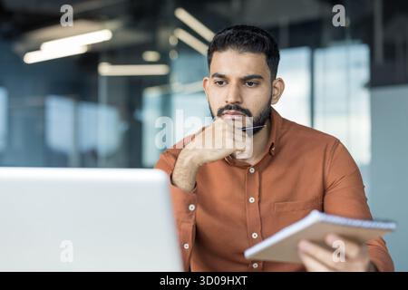 Indian man deeply concentrating while reviewing content on his laptop and holding a pen and notepad, reflecting business strategy or problem-solving in a modern office environment Stock Photo