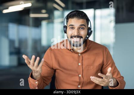 Young man wearing a headset and smiling, offering customer service assistance or participating in an online meeting, communicating with clients virtually from an office Stock Photo