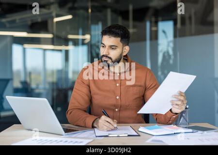 Young man working at a desk, reviewing documents and signing papers while using a laptop in a contemporary office environment, symbolizing business management and professional productivity Stock Photo