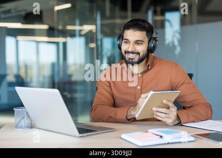 Smiling young man in an open office setting wearing headphones, actively participating in a virtual meeting or online course on a laptop while taking notes in a notebook Stock Photo
