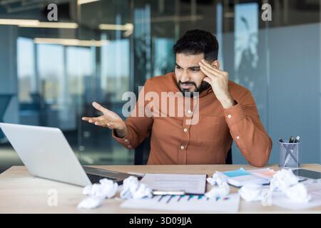 Frustrated young man experiencing professional burnout and headache, having problems with work, looking distraught at his laptop with crumpled papers on the office desk Stock Photo