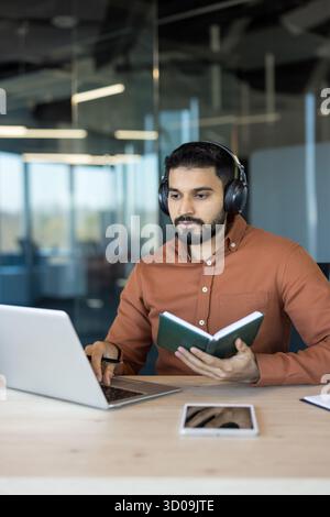 Young indian man wearing headphones and concentrating on his laptop, actively participating in a virtual meeting or e-learning session while holding a notebook and pen in a modern office setup Stock Photo
