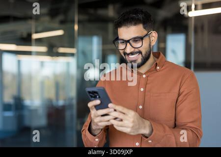 Happy young bearded indian man in glasses smiling while messaging on mobile phone, enjoying communicating and connecting with friends or colleagues in a corporate office setting Stock Photo
