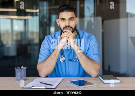 Young bearded man doctor wearing blue scrubs and stethoscope, sitting at desk with papers and tablet, contemplating medical cases in a contemporary office setting Stock Photo
