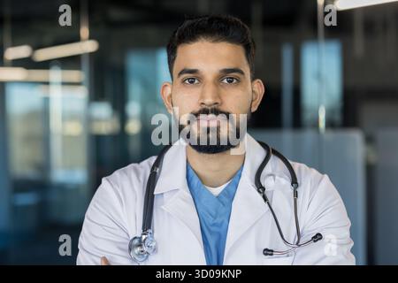 Young indian male physician wearing a white lab coat and stethoscope, standing confidently in a modern medical office setting, representing healthcare professionalism and expertise Stock Photo