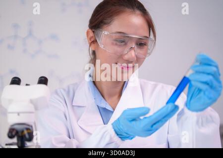 Asian woman professional performs chemical analysis She wears protective gear observing a vibrant blue solution in glass tube This image conveys medic Stock Photo