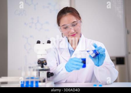 Asian woman adult researcher performs chemical test using lab equipment in modern scientific facility investigating blue solution diligently. She wear Stock Photo