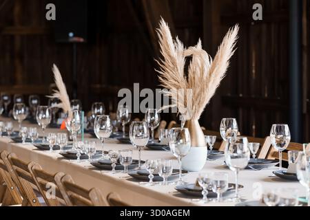 A beautifully arranged long table is set for a wedding reception inside a rustic wooden barn, featuring pampas grass in vases as centerpieces and eleg Stock Photo