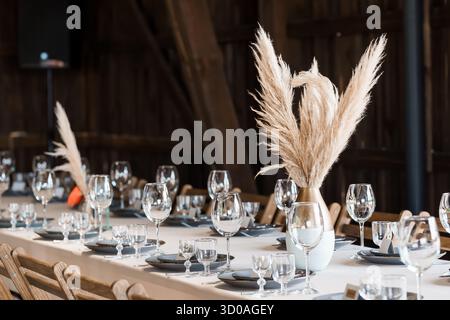 A beautifully arranged wedding reception table features delicate glassware, dark plates, and striking pampas grass in vases, set within a charming rus Stock Photo
