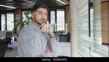 Leaning man wearing light blue shirt and watch near column in open-plan office, with code overlay Stock Photo