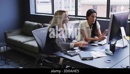 Collaborating women wearing blouse and blazer pointing at PC screen on office desk, with tablet Stock Photo