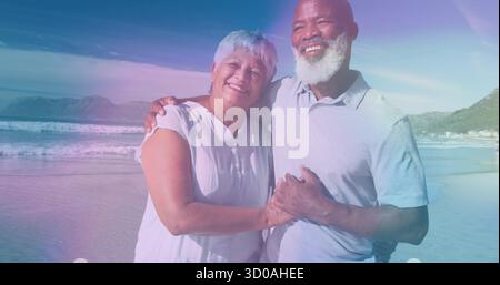 Smiling bearded Black senior man hugging grey-haired Hispanic woman at sandy beach with ocean glow Stock Photo