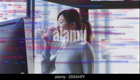 Holding Asian developer drinking coffee from reusable cup by monitor at office, with code overlay Stock Photo