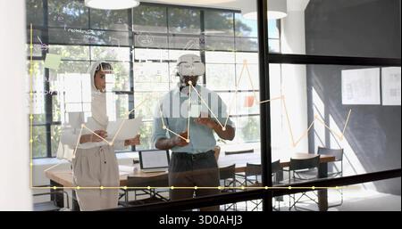 Collaborating team using VR headset and marking diagrams on glass wall in office, with laptops Stock Photo