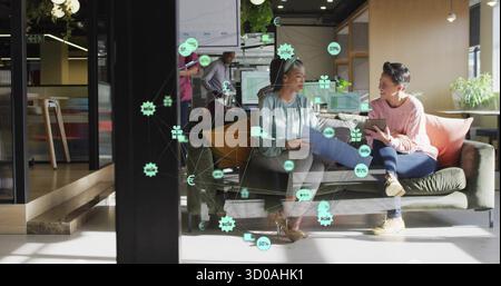 Discussing two women on sofa analyzing charts and data overlays in office lounge, with tablets Stock Photo