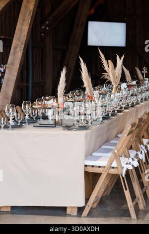 A beautifully arranged wedding reception table inside a rustic barn features a long beige linen cloth, elegant glassware, and pampas grass centerpiece Stock Photo